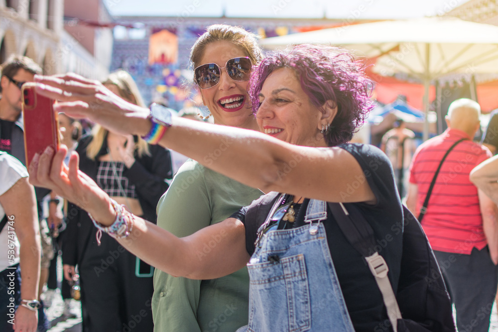 Fototapeta premium Happy mature friends taking a photo. Cheerful mature friends laughing in casual clothes at a street gift market take a selfie photo on cell phone in the crowd.