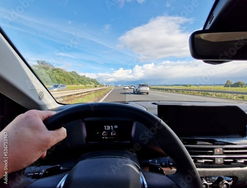 Driver point of view from car interior while driving a car on highway. Drivers hand holding steering wheel and driving on highway during day. POV shot