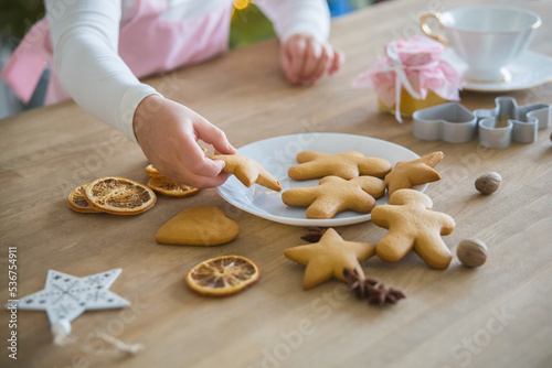 A child's hand takes from a white plate Christmas homemade cookies in the shape of a star, On a wooden table lie cookies gingerbread man, dried orange, anise, peenium molds and a white forfor cup