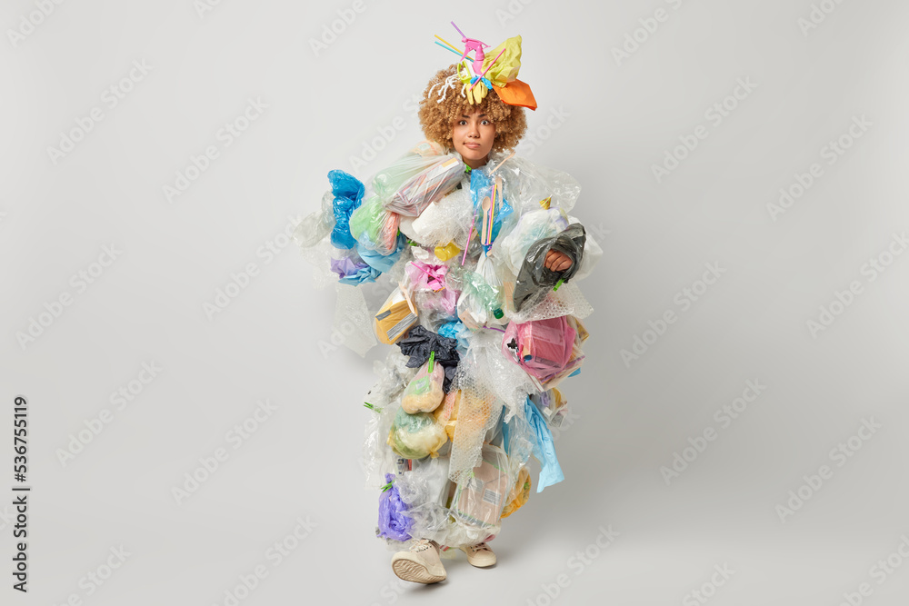 Full length shot of displeased curly haired woman dressed in rubbish costume takes care of environmental protection walks indoor against grey studio background. Ecology and pollution concept