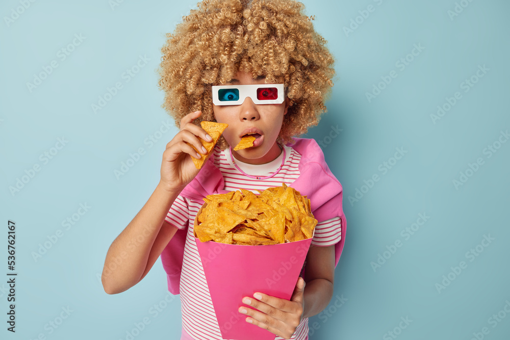 Scared woman bites crisps reacts to something horrible holds breath from amazement wears cinema glasses watches horror movie dressed in striped t shirt sweater tied over shoulders blue background Scared woman bites crisps reacts to something horrible holds breath from amazement wears cinema glasses watches horror movie dressed in striped t shirt sweater tied over shoulders blue background