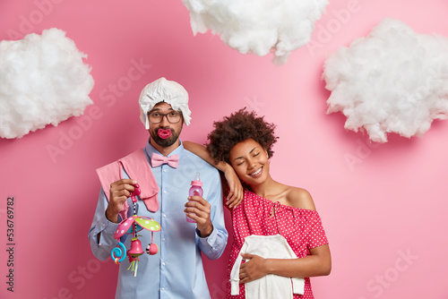 Upset man and his pregnant wife prepars for child birth going to become parents pose with baby supplies isolated over pink background with white clouds overhead. Family and parenthood concept photo