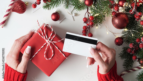Hands of woman in red sweater holding credit card and gift box on white background close-up,top view,flat lay,copy space.Christmas and New Year online shopping,credit card payment.Buying gifts online.
