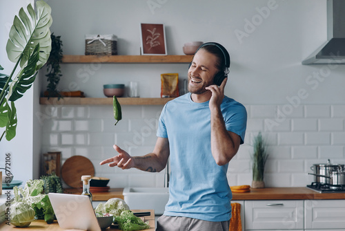 Playful young man in headphones tossing up a green pepper while preparing food at the kitchen