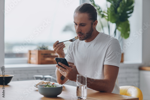 Young man enjoying lunch and using smart phone while sitting at the kitchen