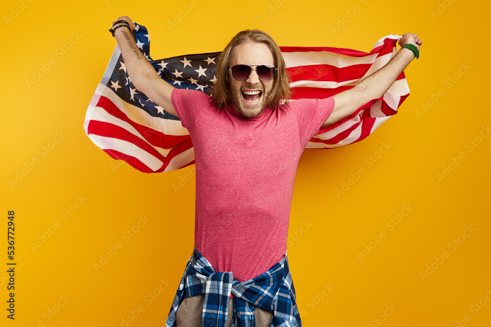 Excited young man carrying American flag while standing against yellow background Excited young man carrying American flag while standing against yellow background