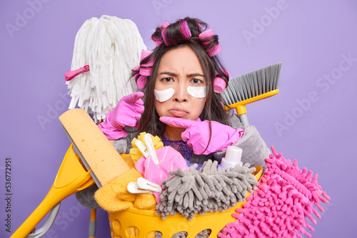 Angry disappointed Asian housewife undergoes beauty procedures and does housework at same time surrounded by cleaning equipment applies hair rollers and beauty patches poses indoors purple wall photo