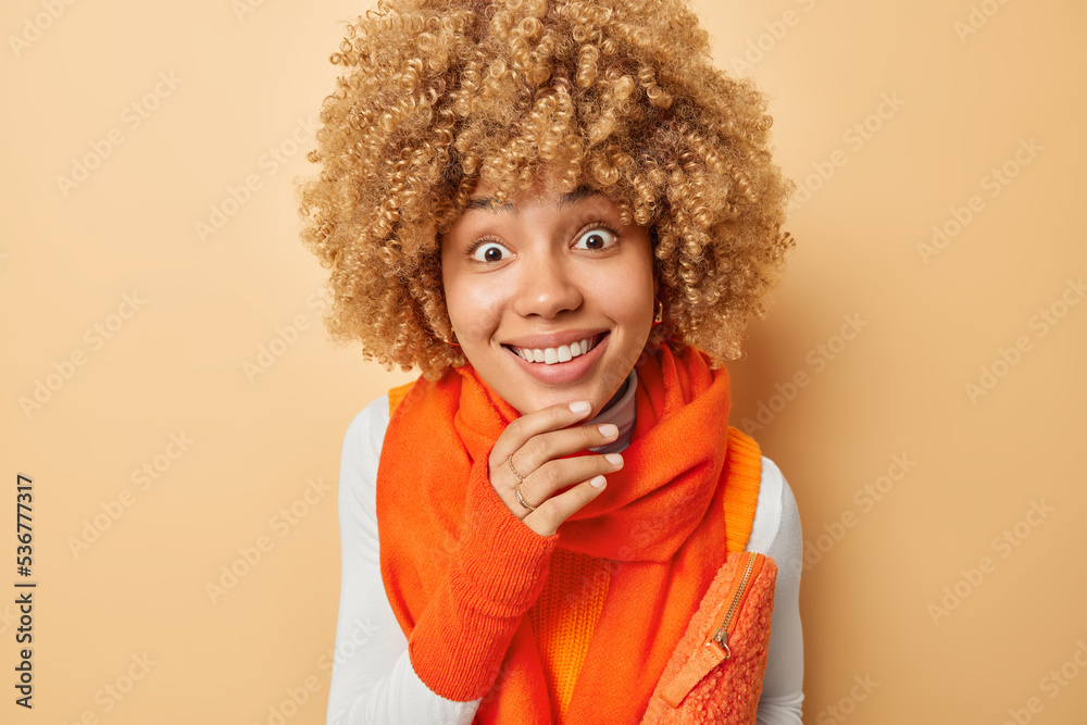 Headshot of positive curious young woman with curly hair keeps hand on chin wears orange scarf and gloves carries bag under arm stares with eyes full of interest isolated over beige background Headshot of positive curious young woman with curly hair keeps hand on chin wears orange scarf and gloves carries bag under arm stares with eyes full of interest isolated over beige background