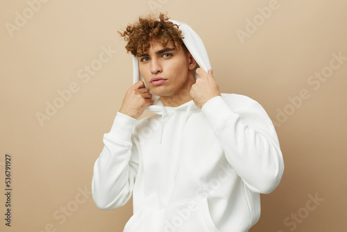 a stylish young man in a white hoodie, standing on a beige background and holding the hood on his head with his hands, smiling pleasantly at the camera