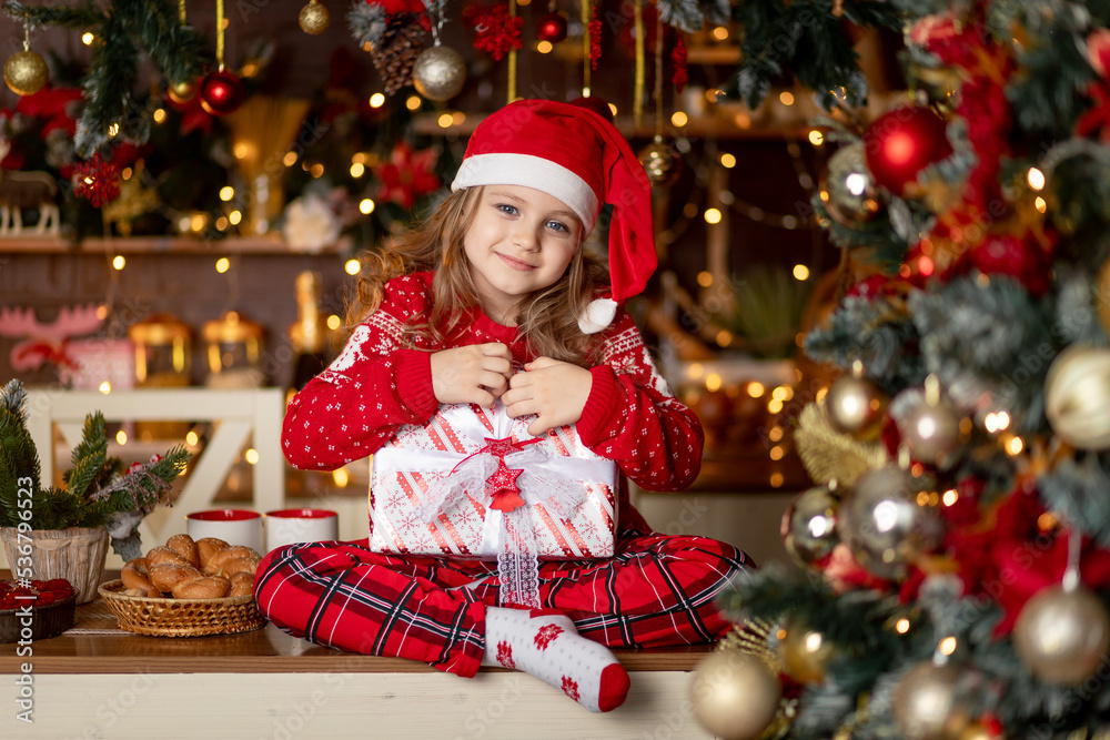 a little cute girl child in a red sweater and a Santa Claus hat in a dark kitchen with a Christmas tree rejoices with gifts and waits for the new year or Christmas a little cute girl child in a red sweater and a Santa Claus hat in a dark kitchen with a Christmas tree rejoices with gifts and waits for the new year or Christmas