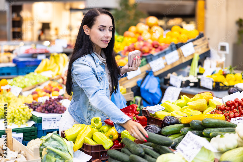 Obraz premium Woman shopping in the afrmers' market reaches out for a cucumber in the vegetables department