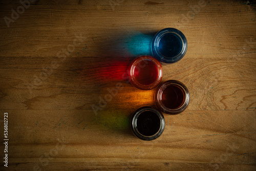 Four glass jars on an old wooden table that contain water with paint and the light reflects the color on the wood