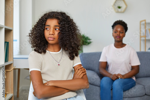 Attractive teenage girl is looking sadly away. Girl sits with her arms crossed in closed pose. Defocused Mom sitting behind and looks at her daughter. African american family at home.