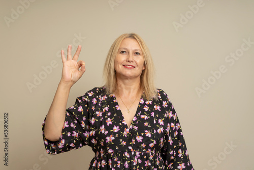 It's okay. Happy woman in patterned dark dress looking approvingly at camera showing ok gesture, like positive sign, approving something good, isolated alone on beige studio background