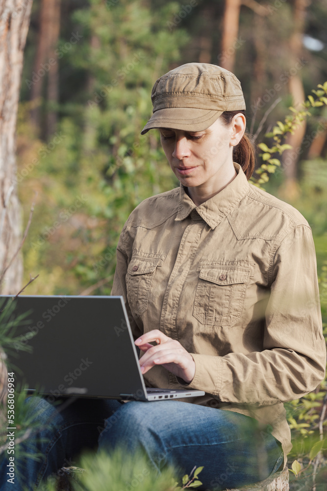 A woman ecologist in uniform sits in a forest area in summer and using a laptop, vertical photo, selective focus.