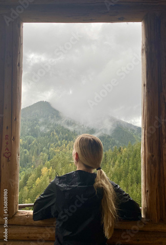 Blond woman looking out of wooden lodge window onto foggy mountain, black forest, Germany