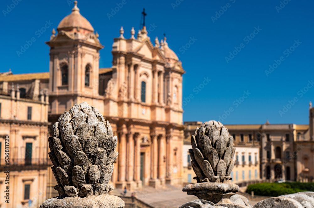 In the foreground baroque decorations in stone in the shape of a pine cone that frame the facade of the cathedral of Noto placed in the background out of focus