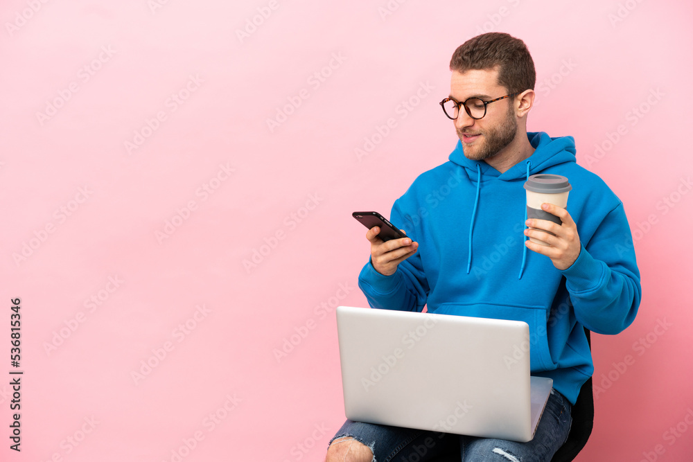 Young man sitting on a chair with laptop holding coffee to take away and a mobile Young man sitting on a chair with laptop holding coffee to take away and a mobile
