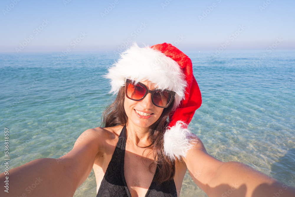 Obraz premium Christmas and holidays concept - Beautiful woman in Santa's hat takes a selfie on the seashore
