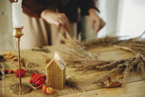 Stylish candles on background of woman hands arranging dried grass in wreath on wooden table. Making stylish autumn wreath on rustic table. Fall decor and arrangement in farmhouse