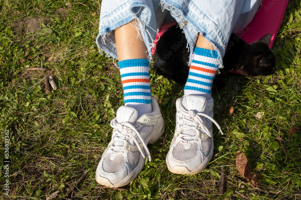 Legs of a girl in stripe bright funny socks and white sneakers. Young woman and black cat sitting on pink mat on green grass. Top view