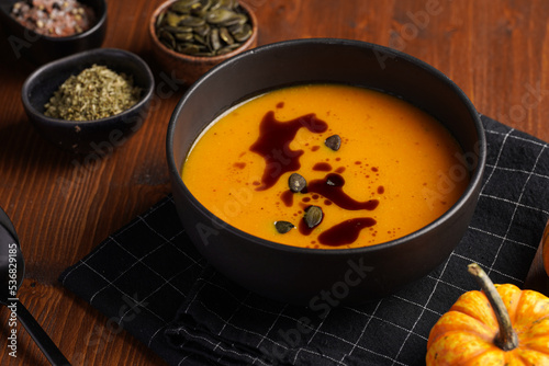 One deep black bowl with an orange pumpkin cream soup with pumpkin seed oil, two black spoons on a wooden surface