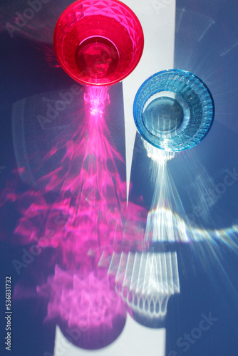 Glittering colored glasses of water on blue background in sunlight. Sunlight through glass of water. Long harsh shadows. Summer vacation concept. Vertical photo.