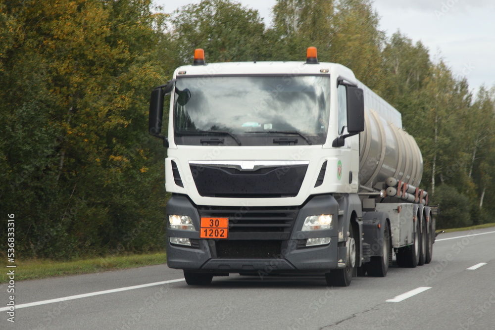 White semi truck barrel truck with 30-1202 dangerous goods class sign drive on suburban highway road at autumn day, front view, diesel fuel ADR cargo transportation logistics