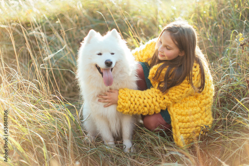 A little brunette girl in a knitted yellow coat with her white fluffy dog lies on autumn leaves in the park. Friendship and harmony in the family. Happy childhood. © viktoriia1974