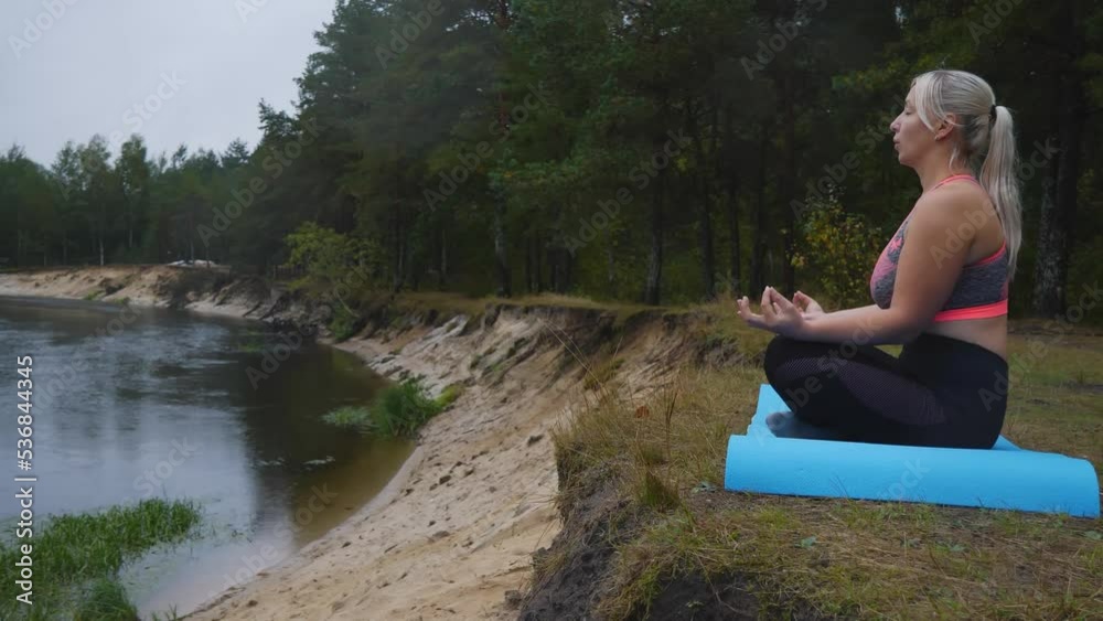 A blonde woman breathes slowly while sitting in a lotus position on the edge of a sandy river bank. In the evening, the woman decided to meditate and do yoga. The concept of meditation near the river. A blonde woman breathes slowly while sitting in a lotus position on the edge of a sandy river bank. In the evening, the woman decided to meditate and do yoga. The concept of meditation near the river.