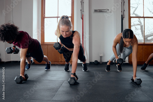 Three determined young adults in a row doing plank exercises with weights at the gym
