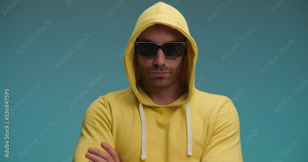 casual young man with yellow sweatshirt nodding, taking sunglasses off and making a funny face on blue background in studio