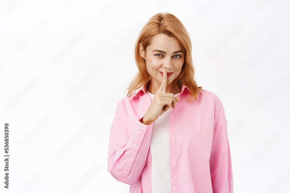 Secret. Smiling young woman showing hush, shush taboo sign, hiding smth, tells to be quiet, stands over white background Secret. Smiling young woman showing hush, shush taboo sign, hiding smth, tells to be quiet, stands over white background