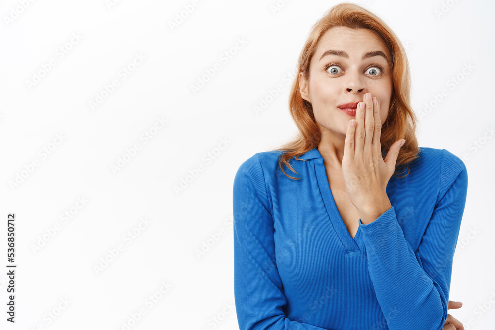 Young woman looks surprised and intrigued by gossips, covers her mouth, talking about smth interesting, standing over white background Young woman looks surprised and intrigued by gossips, covers her mouth, talking about smth interesting, standing over white background