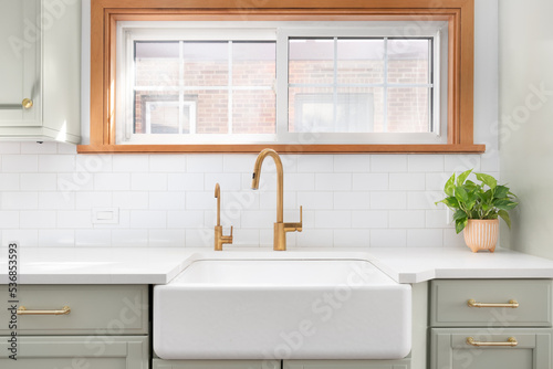 A kitchen sink detail shot with a gold faucet  farmhouse sink  subway tile backsplash  and a light green cabinet.