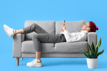 Pretty young woman with unusual hair and phone lying on sofa against blue background
