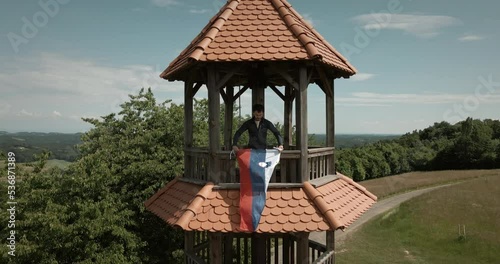 Drone aerial shot of a hiker man hanging Slovenian flag over fence on a small tower on a top of the hill in nature with a forest and agricultural fields around in Slovenia, Sotinski breg photo