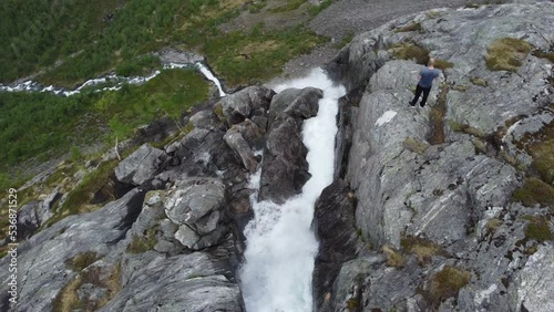 Tourist standing on edge of steep and dangerous mountain cliff with waterfall Fjellfossen falling down - Aerial flying over edge and looking far down in valley below - Eidslandet Stamnes Norway photo