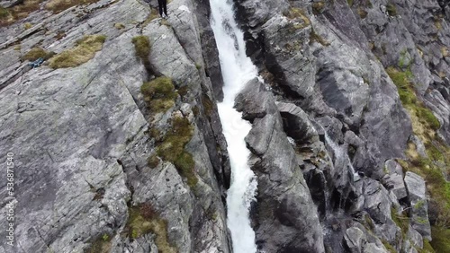 Rotating and backward moving aerial showing tourist standing on edge of steep cliff while operating drone to show amazing waterfall Fjellfossen - Eidslandet Norway aerial photo