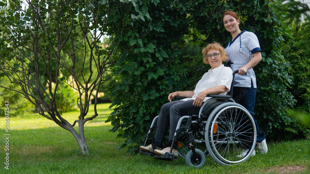 Caucasian female doctor walks with an elderly patient in a wheelchair in the park. Nurse accompanies an old woman on a walk outdoors. Caucasian female doctor walks with an elderly patient in a wheelchair in the park. Nurse accompanies an old woman on a walk outdoors.