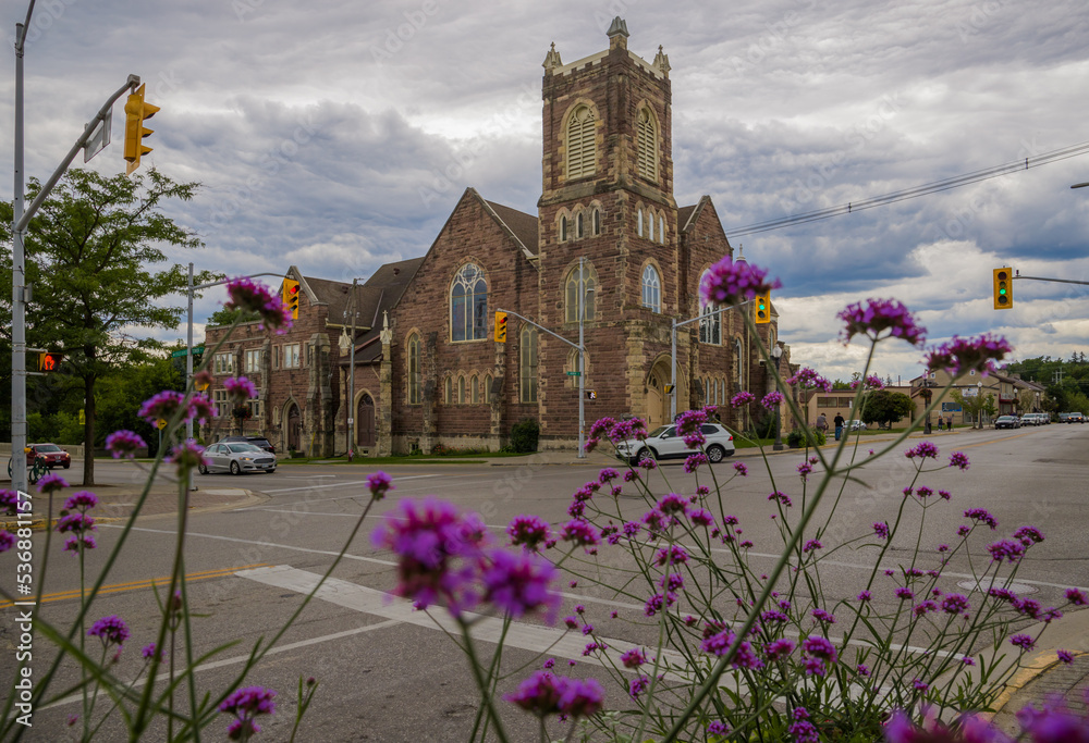 Old Catholic stone church at crossroads in downtown Fergus, Ontario, Canada. Cloudy sky, violet flowers