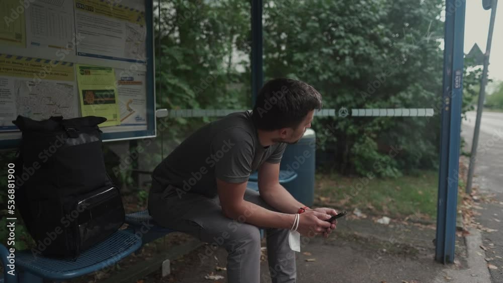 Caucasian man waits long time for bus at bus stop, is annoyed and looks at his watch and looks out at road. A passenger at the bus stop is surfing the Internet on his smartphone. Munich. Germany. 