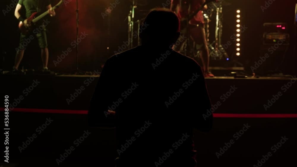 unrecognizable Sound Engineer adjusts sound for rock concert in large event hall. Rear view silhouette of man in spotlights with artists and guitarists on stage in background. unrecognizable Sound Engineer adjusts sound for rock concert in large event hall. Rear view silhouette of man in spotlights with artists and guitarists on stage in background.