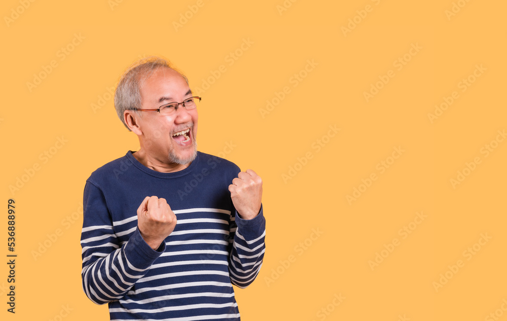Yes! Portrait of Asian senior man wearing glasses standing raising his fists and excited facial expression isolated on yellow background. Yes! Portrait of Asian senior man wearing glasses standing raising his fists and excited facial expression isolated on yellow background.