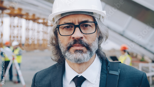 Fotografia Close-upportrait of serious adult man investor standing at industrial site wearing helmet and looking at camera