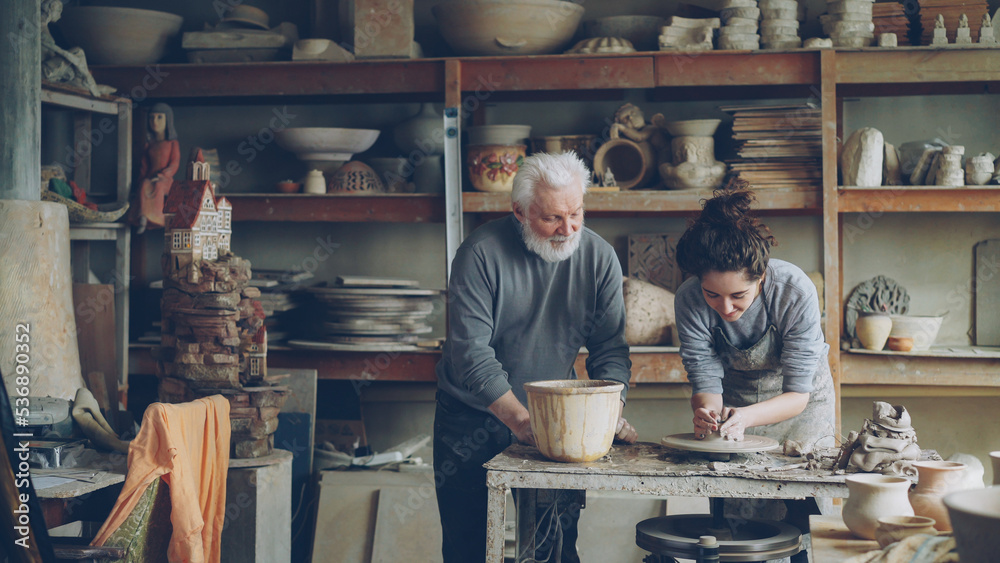Caring senior grandfather is showing young granddoughter how to work with clay on throwing-wheel in small workshop. Pottery, family hobby and handicraft concept.