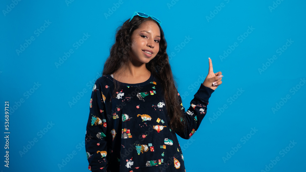 Niña con fondo azul usando una pijama de navidad,
posando con lentes y señalando con el dedo Niña con fondo azul usando una pijama de navidad,
posando con lentes y señalando con el dedo