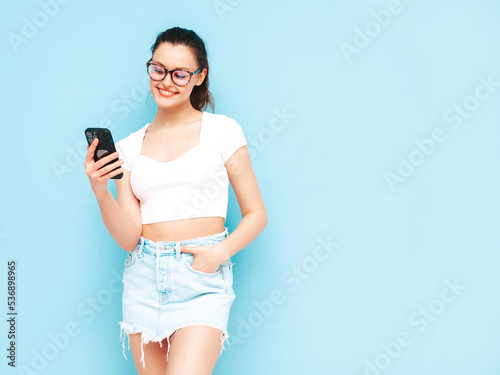 Young beautiful smiling female in trendy summer jeans skirt. Sexy carefree woman posing near blue wall in studio. Positive brunette model looking at cellphone screen. Holding smartphone and using apps