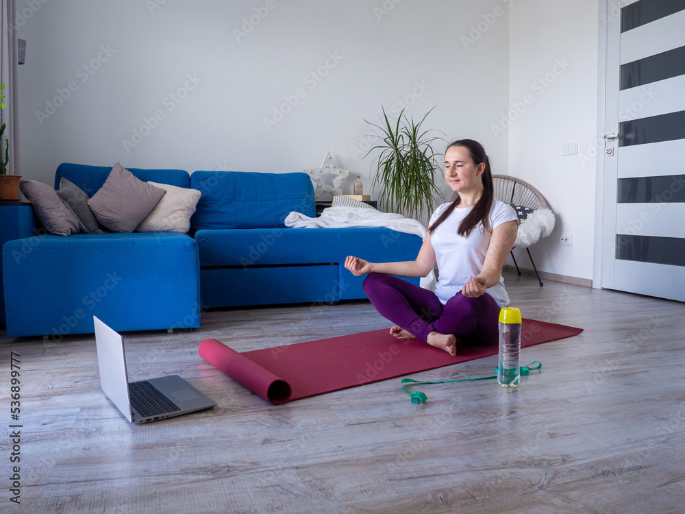 European girl looking on laptop during practising yoga. Concept of fitness, sport at home and healthy lifestyle. Online training, watching video tutorial on a laptop. Woman doing exercises on yoga mat
