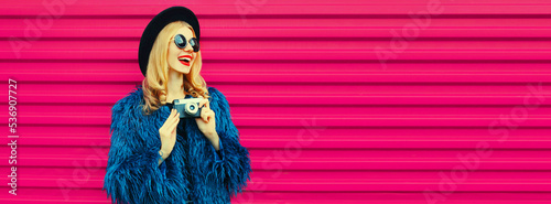 Portrait of stylish happy smiling young woman photographer with film camera taking picture wearing blue fur coat, black round hat on pink background © rohappy